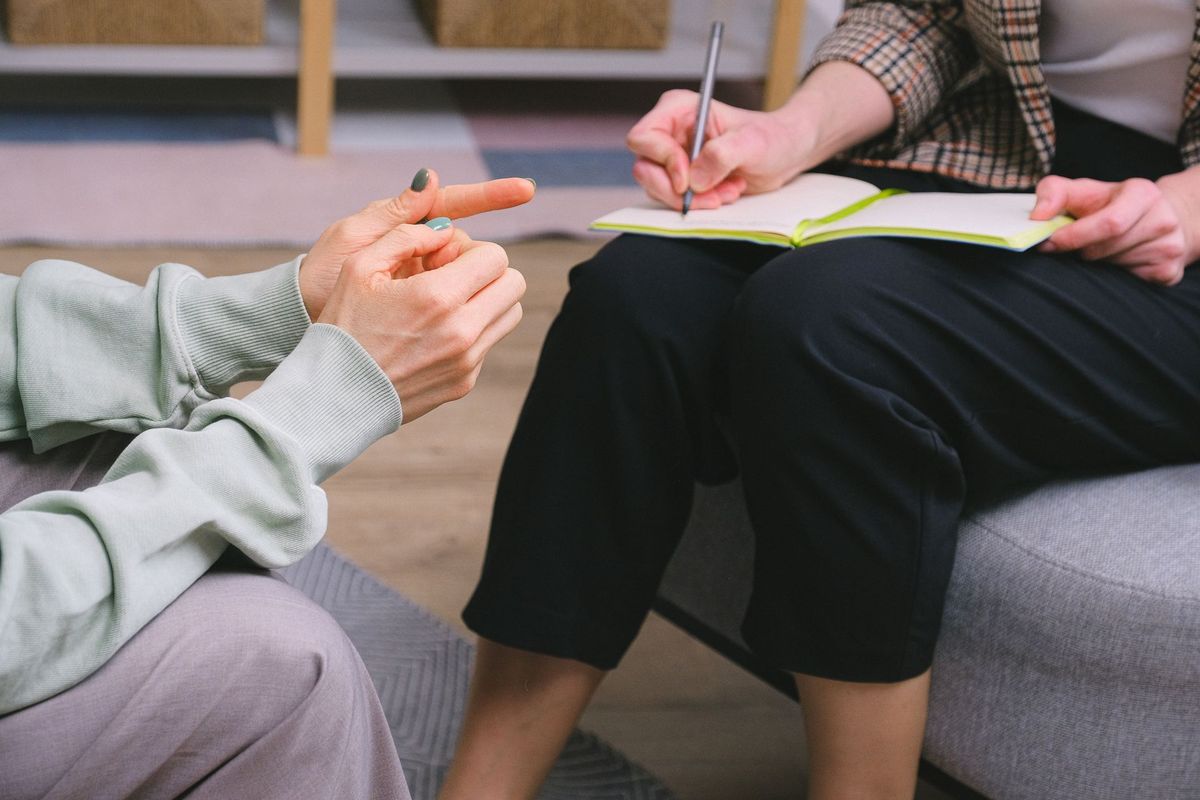 A professional takes notes in an open notebook while the person seated across from them gestures as they talk.