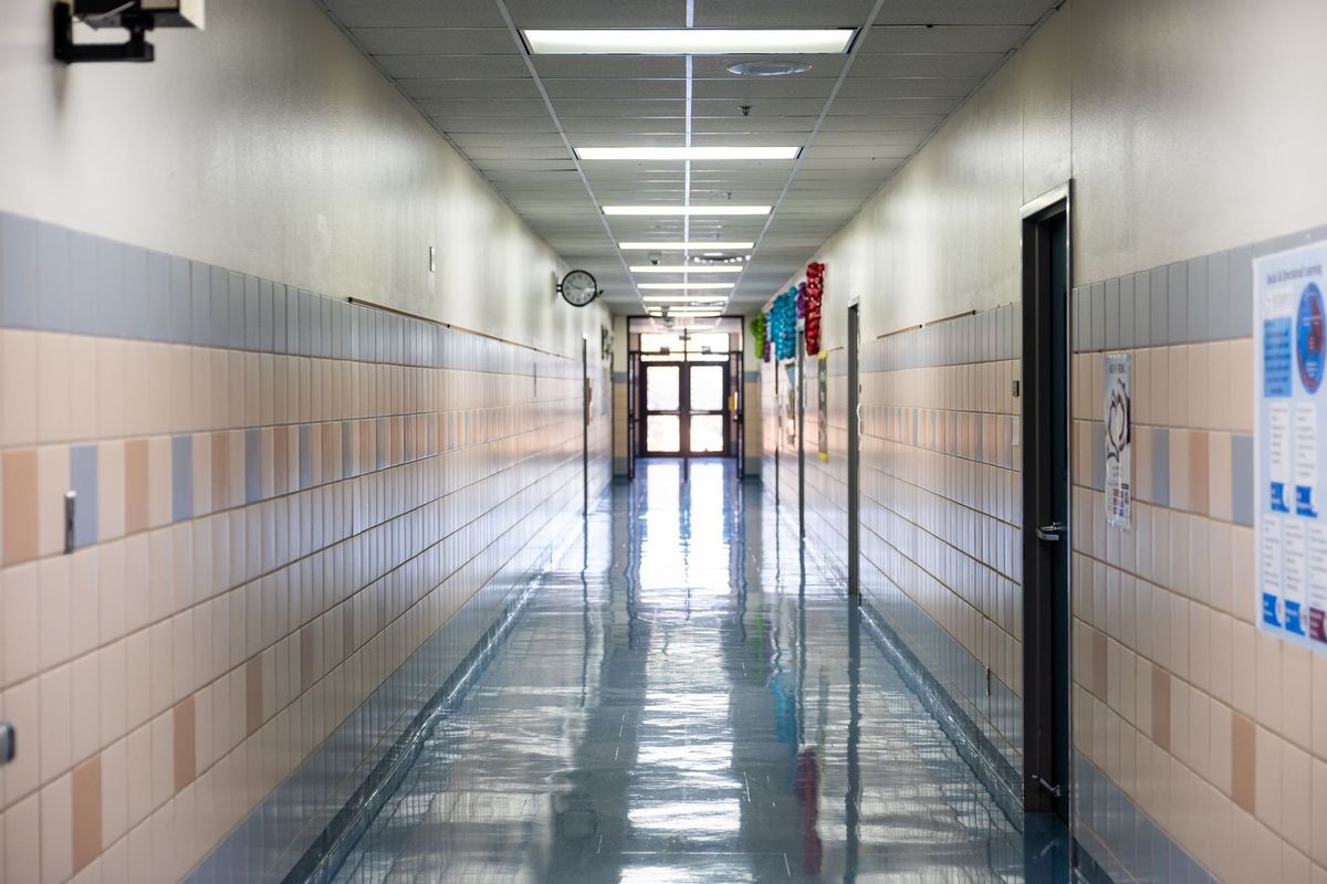 An empty school corridor with tiled walls and a polished floor, leading to glass double doors at the far end.