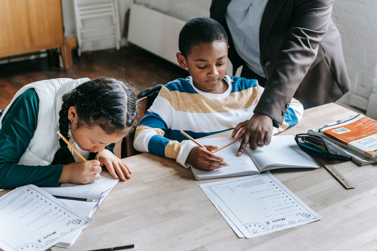 A teacher points to a boy's notebook as he and a classmate work through math worksheets at a shared classroom table.